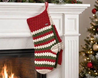 May include: A handmade Christmas stocking in red, green, and white stripes hangs from a white mantel. The stocking is crocheted with a loop for hanging. A Christmas tree and fireplace are in the background.