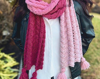 May include: A handmade crochet scarf in shades of pink and burgundy. The scarf features a textured design and is adorned with tassels at the ends. The Crochet Village logo is visible in the bottom left corner.