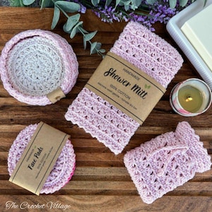 May include: A collection of handmade, light pink crochet bath accessories, including a round coaster, a washcloth, a shower mitt, and face pads. Each item has a label with the text "HANDMADE" and "100% COTTON". The items are arranged on a wooden surface.