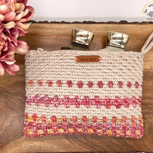 Può includere: Una pochette con cerniera in beige all'uncinetto con un'etichetta in pelle marrone "Hand Made". La pochette ha un design sfumato rosa, arancione e giallo nella parte inferiore e una striscia rosa al centro. La pochette è aperta e contiene due bottiglie di vetro trasparente con tappi argentati.