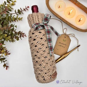 May include: A beige crochet bottle cover featuring a textured cable knit pattern and a red, green, and white plaid ribbon tied in a bow around the neck. A brown paper tag with the text Handmade with love is attached by a string next to a gold hook.