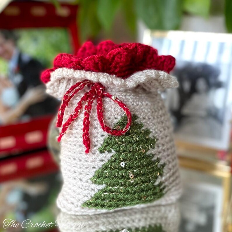 May include: A crocheted Christmas gift bag with a red drawstring and a green Christmas tree design. The bag is decorated with gold sequins.