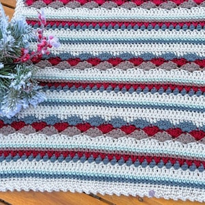 May include: A crocheted blanket featuring horizontal stripes in cream, light blue, dark gray, and deep red. The pattern includes rows of textured shell stitches and solid stitches. A sprig of frosted pine needles with red berries is placed on the left side.