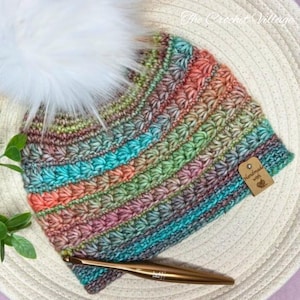 May include: A handmade crochet hat with a fluffy white pom-pom. The hat features a colorful striped pattern in shades of teal, orange, green, and purple. A small tag reads "Handmade with love." A gold crochet hook is also visible.