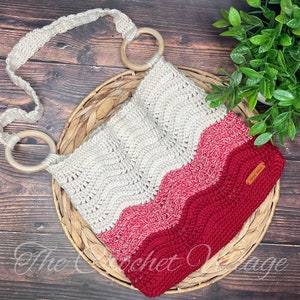Peut inclure: Un sac en crochet avec un motif ondulé dans les tons de blanc, rose et rouge. Le sac a deux anneaux en bois sur la bandoulière et une petite étiquette en cuir marron avec le texte "The Crochet Village".