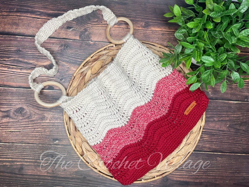 May include: A crocheted bag with a wavy pattern in shades of white, pink, and red. The bag has two wooden rings on the strap and a small brown leather tag with the text "The Crochet Village".