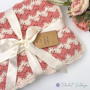 May include: A folded crochet blanket featuring a repeating shell stitch pattern in dusty rose and cream yarn. The blanket is tied with a wide cream satin ribbon and includes a brown rectangular gift tag printed with "Handmade with love! From".