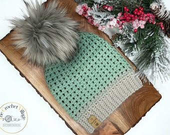 May include: A green and beige crocheted beanie with a grey faux fur pom pom. The beanie is on a wooden surface with a tag that says "Handmade with Love".