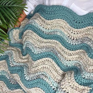 May include: A handmade crochet blanket in shades of teal, gray, and beige. The blanket features a wavy pattern, with the colors alternating in bands. The texture of the yarn is visible, giving the blanket a cozy appearance. The blanket is folded.
