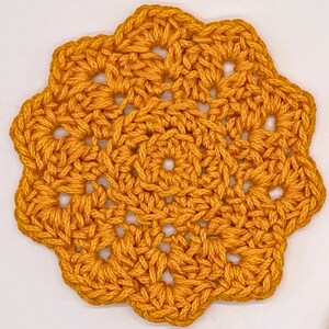 May include: A circular, mustard yellow crochet coaster featuring an intricate, openwork floral pattern with a central ring and eight distinct, scalloped petal-like edges radiating outwards, crafted from thick, textured yarn in a repeating stitch design.