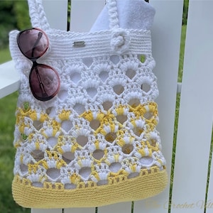May include: White crochet tote bag with a yellow crocheted bottom. The bag has a flower design and is hanging on a white wooden chair. Sunglasses are hanging from the bag.