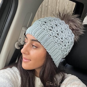 Op de afbeelding: Een lichtblauwe gehaakte muts met een imitatiebont pompon. De muts heeft een textuursteekpatroon en een omgeslagen rand. Het logo van The Crochet Village is zichtbaar. Een stijlvol accessoire voor de winter.