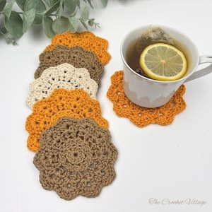May include: A set of six round, scalloped-edge crochet coasters in shades of mustard yellow, brown, and cream. One coaster sits under a white ceramic mug containing tea with a lemon slice and a tea bag. The text The Crochet Village is visible.