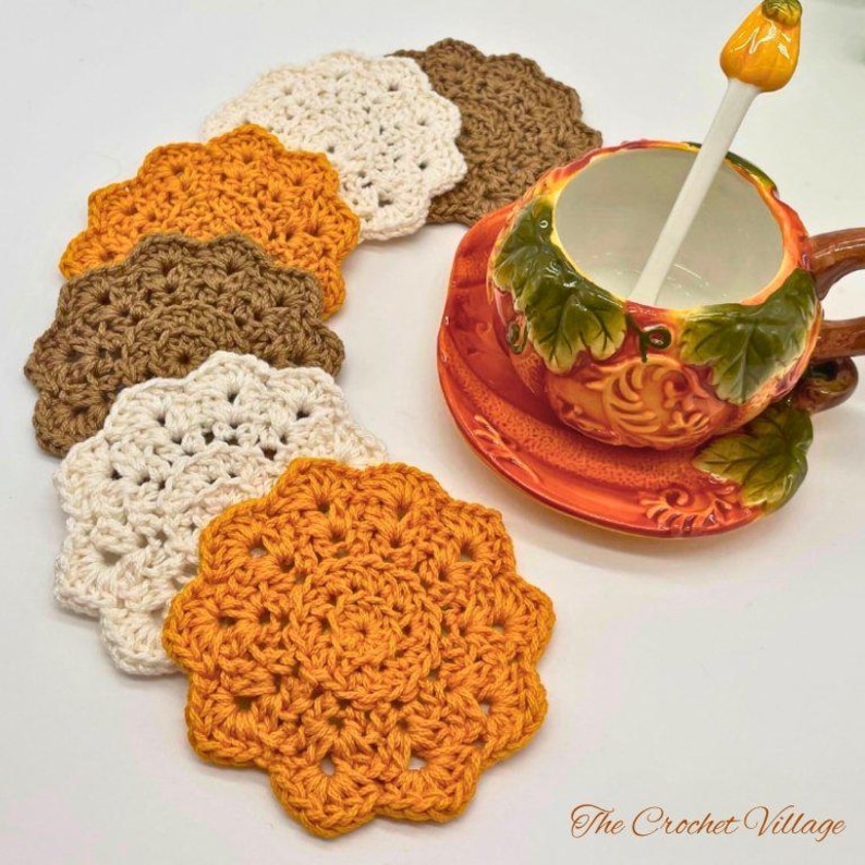 May include: A set of six round, scalloped-edge crochet coasters in cream, orange, and brown, arranged in a curved line. Beside them sits a ceramic pumpkin-shaped teacup and saucer with a white spoon. The text The Crochet Village is visible.
