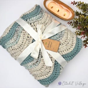 May include: A folded crochet blanket featuring a wavy pattern in alternating shades of cream, light gray, and muted teal. The blanket is tied with a white satin ribbon and includes a brown paper tag with the text Handmade with love.