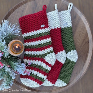 Op de afbeelding: Drie handgemaakte kerstsokken in rood, groen en wit haakwerk. Eén sok heeft horizontale strepen, terwijl de andere effen kleuren hebben met witte accenten. De sokken worden gepresenteerd op een houten dienblad met een brandende kaars en groen.