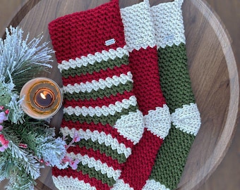 May include: Three handmade Christmas stockings in red, green, and white crochet. One stocking has horizontal stripes, while the others have solid colors with white accents. The stockings are displayed on a wooden tray with a lit candle and greenery.