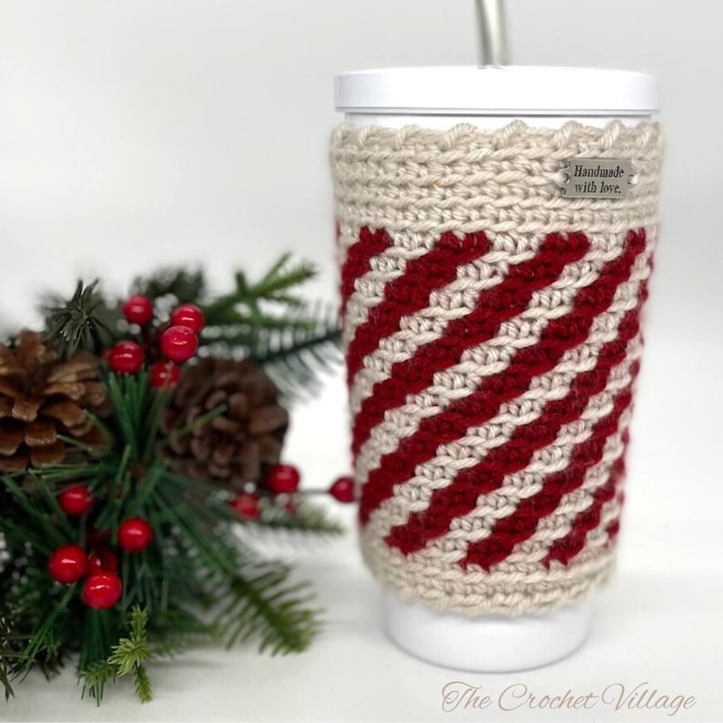 May include: A crocheted cup cozy in red and beige with a diagonal stripe pattern. The cozy has a tag that says 'Handmade with love.' The cozy is on a white cup with a straw.