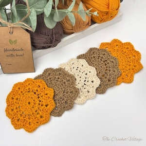 May include: A set of five round, scalloped-edge crochet coasters in shades of mustard yellow, brown, and cream. A brown paper tag with the text "Handmade with love!" is attached to the coasters, which are arranged near balls of yarn.