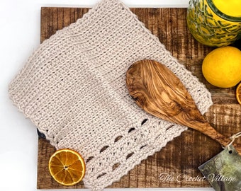 Puede incluir: Un paño de cocina beige tejido a ganchillo con un borde festoneado decorativo, sobre una tabla de cortar de madera. También se ven una cuchara de madera de olivo y una rodaja de naranja seca. The Crochet Village está impreso en una etiqueta.