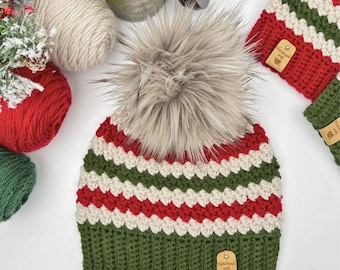 May include: A crocheted beanie featuring horizontal stripes in red, white, and green with a large, fluffy, light brown faux fur pom-pom on top. The hat has a solid green ribbed brim and a small, rectangular, tan-colored tag labeled Handmade with love.