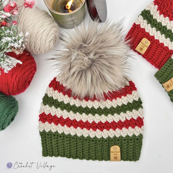 May include: A crocheted beanie featuring horizontal stripes in red, white, and green with a large, fluffy, light brown faux fur pom-pom on top. The hat has a solid green ribbed brim and a small, rectangular, tan-colored tag labeled Handmade with love.