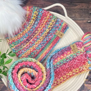 May include: A colourful crocheted hat and scarf set with a white pom pom on the hat. The hat and scarf are made with a multi-coloured yarn with shades of pink, yellow, blue, and green. The set is displayed on a white crocheted surface.