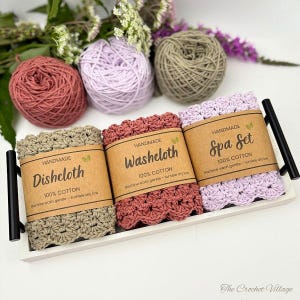 May include: Three crocheted cotton cloths in sage green, dusty rose, and light purple, each wrapped in a brown paper band. The bands are labeled Handmade Dishcloth, Washcloth, and Spa Set, with 100% Cotton and care instructions printed on them.