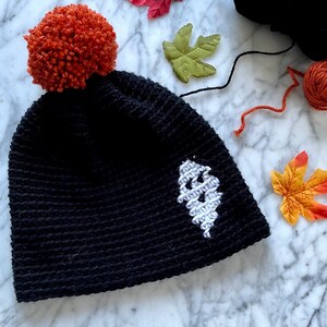 May include: Black crocheted beanie with a white ghost design and a bright orange pom-pom on top.