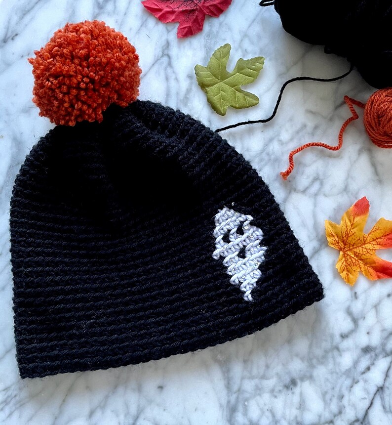 May include: Black crocheted beanie with a white ghost design and a bright orange pom-pom on top.