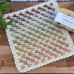May include: A crocheted dishcloth with a scalloped edge in a multi-colored striped pattern. The dishcloth is made with a variety of shades of brown, green, and white yarn. A bar of soap with a label that reads "Dear Botanicals Terrazzo Handmade Artisan Soap Net Wt. 4 oz (113 g)" is sitting on top of the dishcloth.