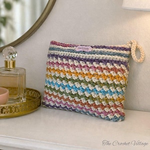 May include: A handmade, colorful crochet pouch with a loop handle. The pouch features a striped pattern in various colors, including purple, yellow, blue, and green. A small tag reads "Handmade". The Crochet Village is written at the bottom.