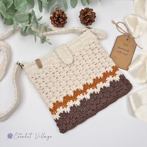 May include: A cream-colored crochet crossbody bag featuring a button closure, a long strap, and horizontal stripes in burnt orange and dark brown at the base. A brown paper tag attached to the bag displays the text Handmade with love.