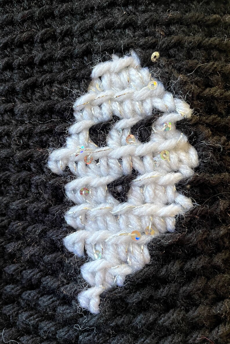 May include: A close-up of a white knitted fabric with silver sequins. The fabric is stitched onto a black knitted fabric.