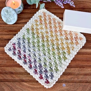 May include: A square crochet washcloth featuring a diagonal gradient pattern of colours including deep red, purple, green, yellow, and orange, bordered by a white edge. The textured fabric is made with a repeating stitch pattern.