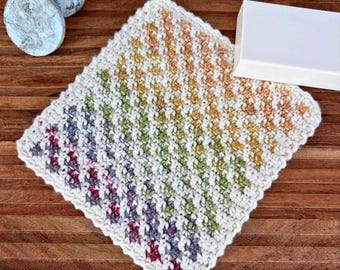 May include: A square crochet washcloth featuring a diagonal gradient pattern of colours including deep red, purple, green, yellow, and orange, bordered by a white edge. The textured fabric is made with a repeating stitch pattern.