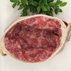 May include: A white crocheted basket with a wooden handle. The basket is lined with a pink fabric with a white floral pattern.