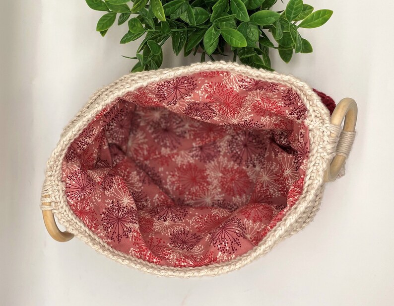 May include: A white crocheted basket with a wooden handle. The basket is lined with a pink fabric with a white floral pattern.