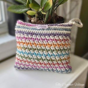 May include: A handmade, colorful crochet plant pot cover. The cover features a textured stitch pattern in a rainbow of colors, including pink, orange, green, and blue. A small tag reads "Handmade". The Crochet Village is written at the bottom.