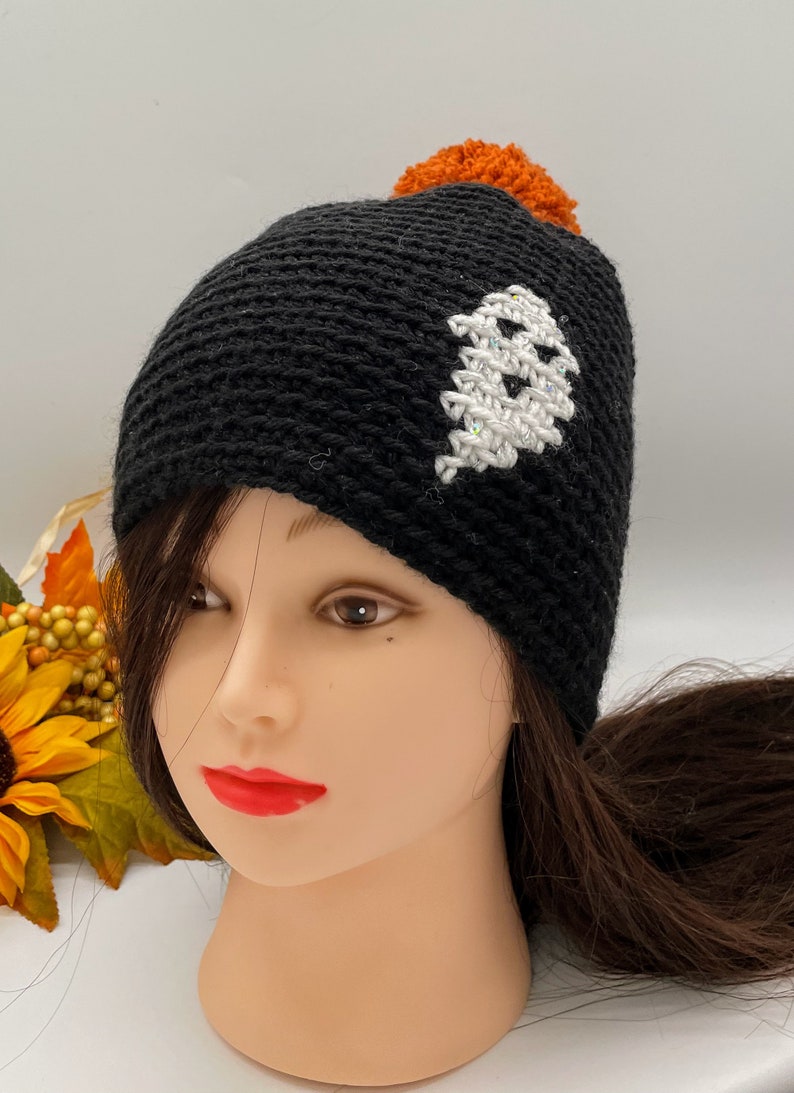 May include: Black crocheted beanie with a white ghost design and an orange pom pom on top.