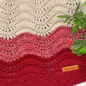 May include: A crocheted blanket with a wavy pattern in shades of beige and red. The blanket has a brown leather tag that says "Hand Made".