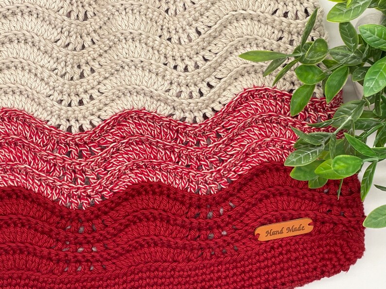 May include: A crocheted blanket with a wavy pattern in shades of beige and red. The blanket has a brown leather tag that says "Hand Made".
