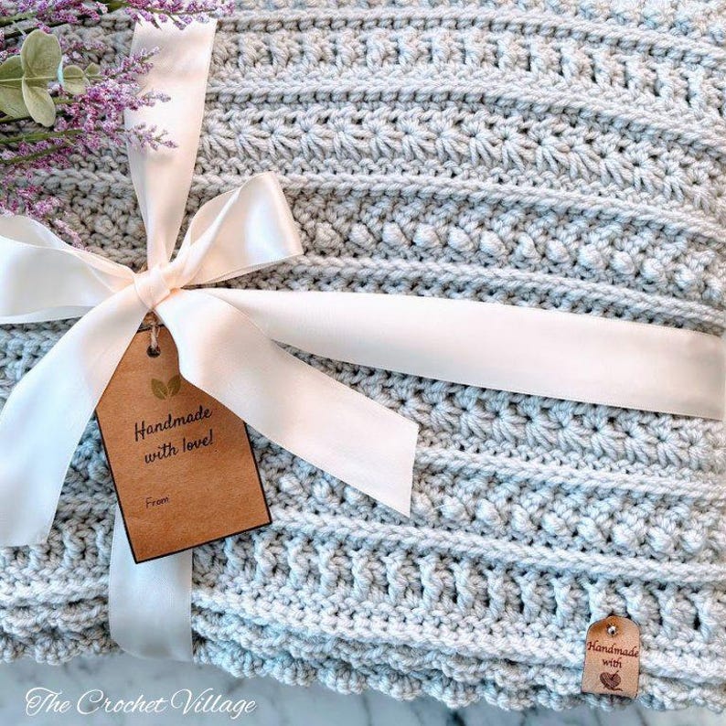 May include: A light gray crocheted blanket featuring horizontal textured rows of stitches. The blanket is tied with a white satin ribbon and includes a brown paper tag printed with the text Handmade with love! and a small wooden label with a heart icon.