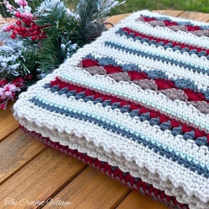 Peut inclure: Une couverture en crochet pliée, de couleur crème, avec un motif répétitif de rayures rouges, grises et bleues. La couverture a un bord festonné et repose sur une surface en bois. Le texte "The Crochet Village" est visible en bas à gauche.