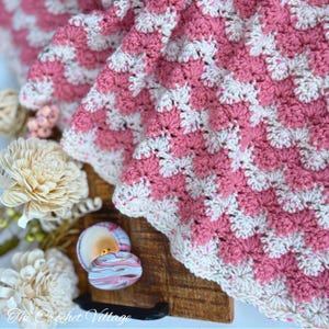 Könnte beinhalten: Handgefertigte Häkeldecke in Rosa und Weiß mit strukturiertem Stichmuster und gewelltem Rand. Das Logo von The Crochet Village ist sichtbar. Die Decke ist auf einer Holzoberfläche drapiert.
