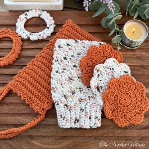 May include: A collection of handmade crochet items in shades of orange and white. The set includes a washcloth, coasters, a hair tie, and a hanging item. The items are arranged on a wooden surface, with a lit candle and greenery in the background.