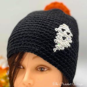May include: Black crochet beanie with a white ghost design and a fluffy orange pom pom on top. The beanie is worn by a person with brown hair and brown eyes. The text "The Crochet Village" is visible at the bottom of the image.