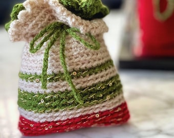 Könnte beinhalten: Eine gehäkelte Geschenktüte mit einem grün-beigen Streifenmuster und rotem Besatz. Die Tasche ist mit goldenen Pailletten verziert.