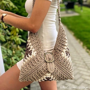 May include: A handmade crochet bag in shades of brown and cream, featuring a geometric pattern and a large buckle closure. The bag has a long strap and is worn over the shoulder. The Crochet Village is printed on the bottom right.