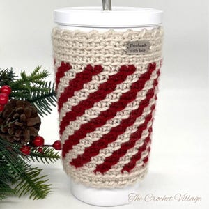 May include: A crocheted cup cozy in red and beige with a "Handmade with love." tag. The cozy is on a white travel mug.